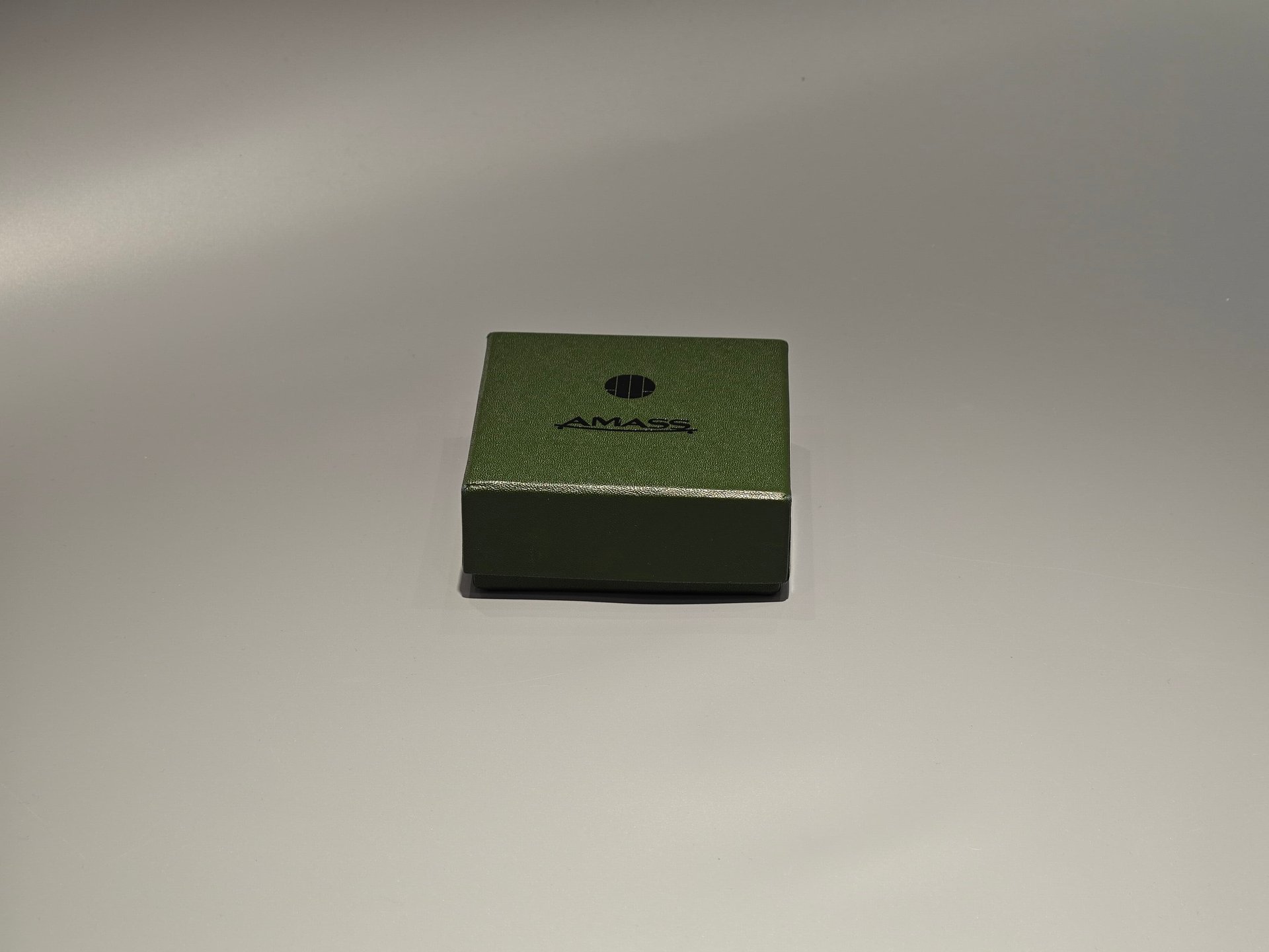 The product is a small green custom box designed with a lift-off lid, suitable for packaging needs. The box features minimalist branding on the front and is set against a plain background.