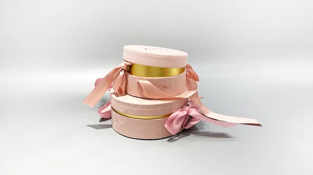 Our product image showcases three custom round gift boxes in a soft pink hue, each embellished with elegant gold bands and topped with neatly tied pink ribbons. These boxes are artfully arranged in a stacked formation against a neutral background, emphasizing the sophisticated design and luxurious appeal of our packaging solutions.